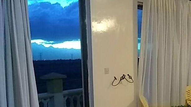 En balkon med udsigt over et bjerglandskab, hvide gardiner, en plastikstol og en lyskontakt på væggen.