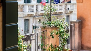 Ein Balkon mit Blumentöpfen, Fliesenboden und ein Gebäude mit mehreren Fenstern sowie Flaggen im Hintergrund.