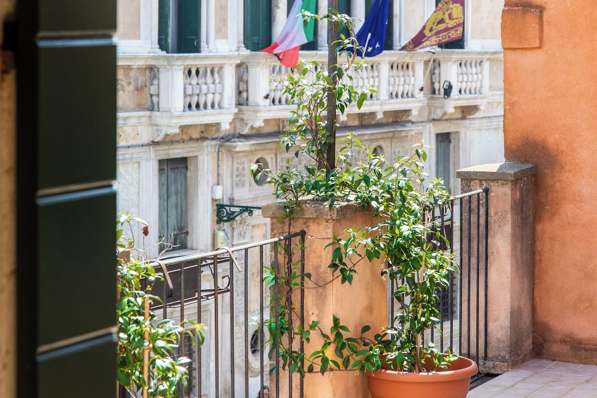 A balcony with potted plants, a tiled floor, and a building with multiple windows and flags in the background.