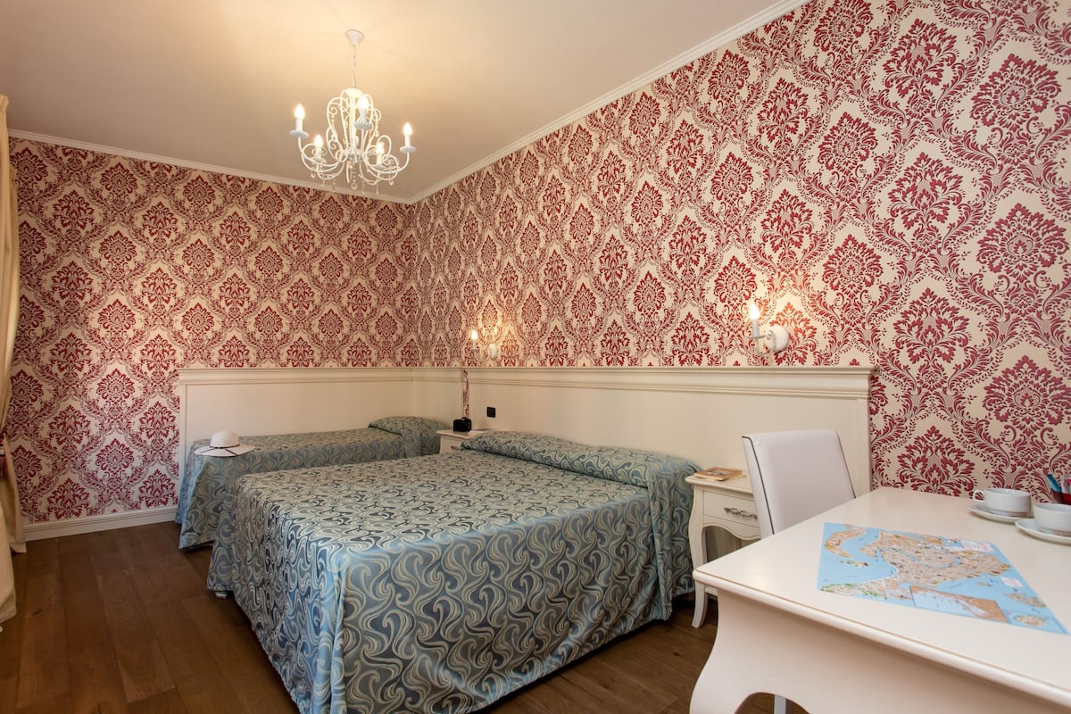 A bedroom with two beds, a chandelier, patterned wallpaper, a desk with a map, and a white chair.