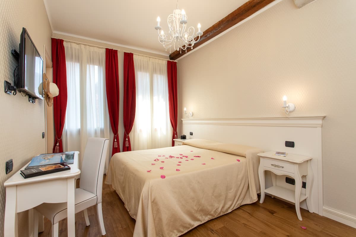A bedroom with a bed, a desk, a chair, a nightstand, a chandelier, and a window with red curtains.