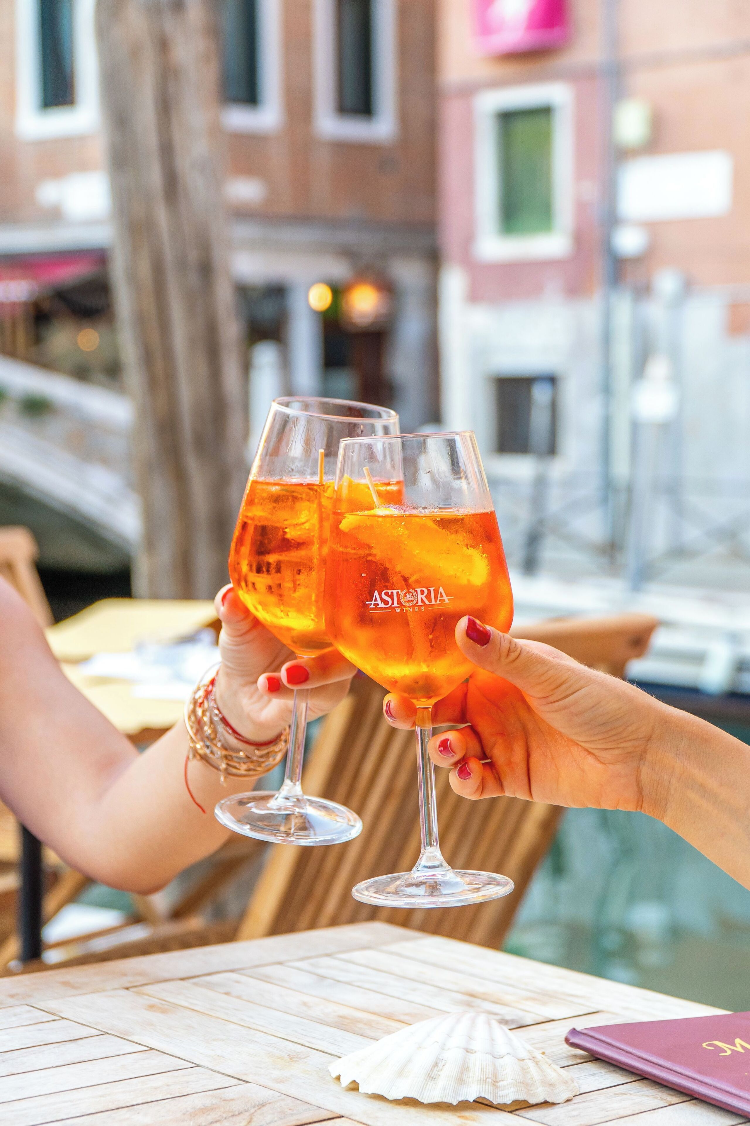 Twee personen proosten met glazen met een goudkleurige drank aan een buitentafel met een houten blad, een schelp en een paars notitieboekje.