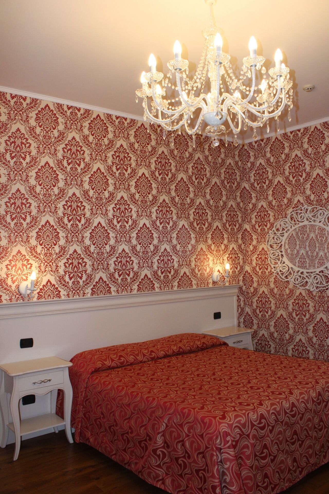 A bedroom with a patterned wallpaper, a chandelier, a bed with a red patterned bedspread, and a nightstand with a drawer.