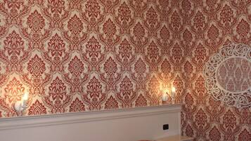 A bedroom with a patterned wallpaper, a chandelier, a bed with a red patterned bedspread, and a nightstand with a drawer.