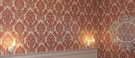 A bedroom with a patterned wallpaper, a chandelier, a bed with a red patterned bedspread, and a nightstand with a drawer.