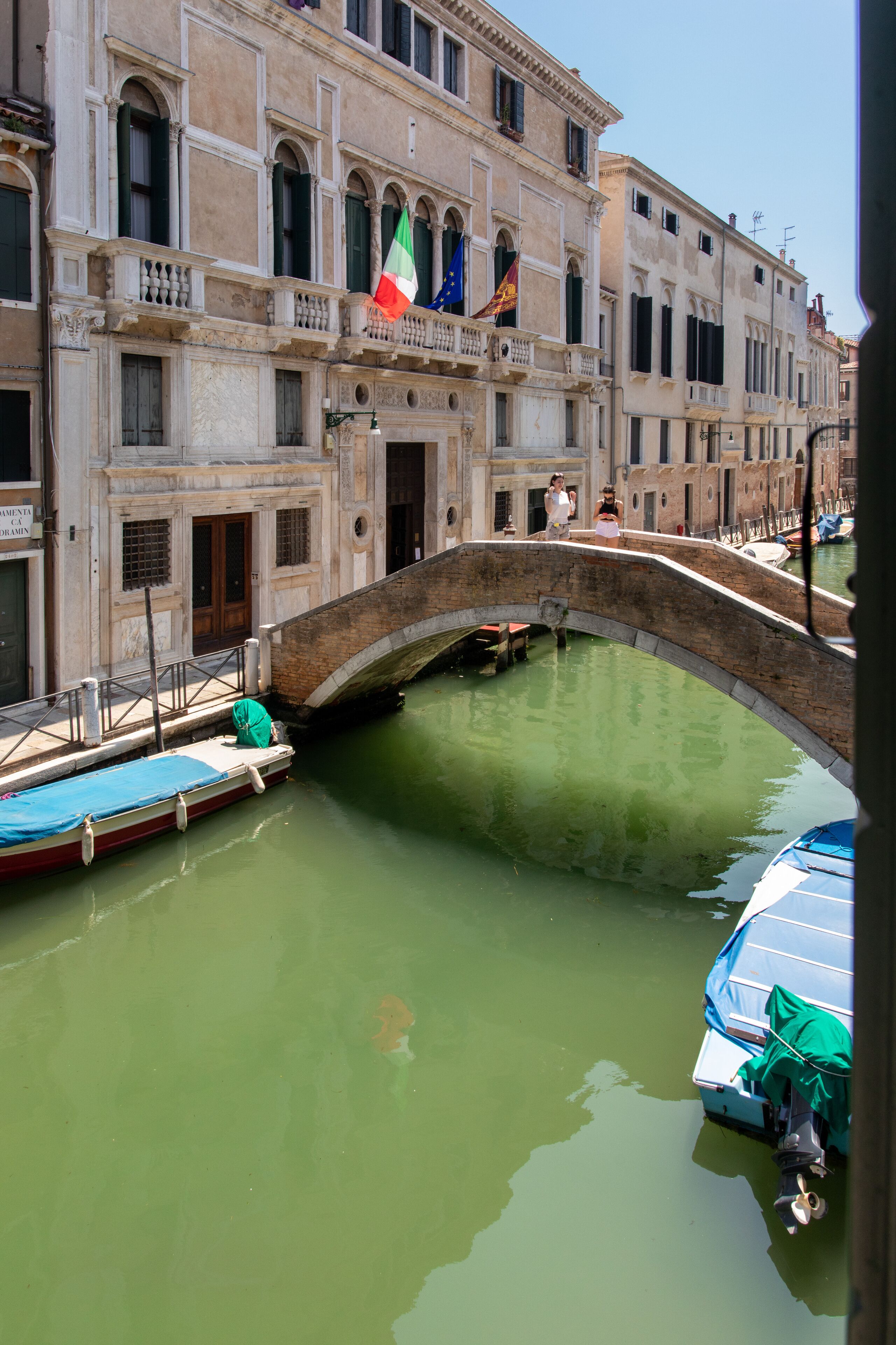 Un canal con un puente de piedra, barcos y edificios históricos con banderas.
