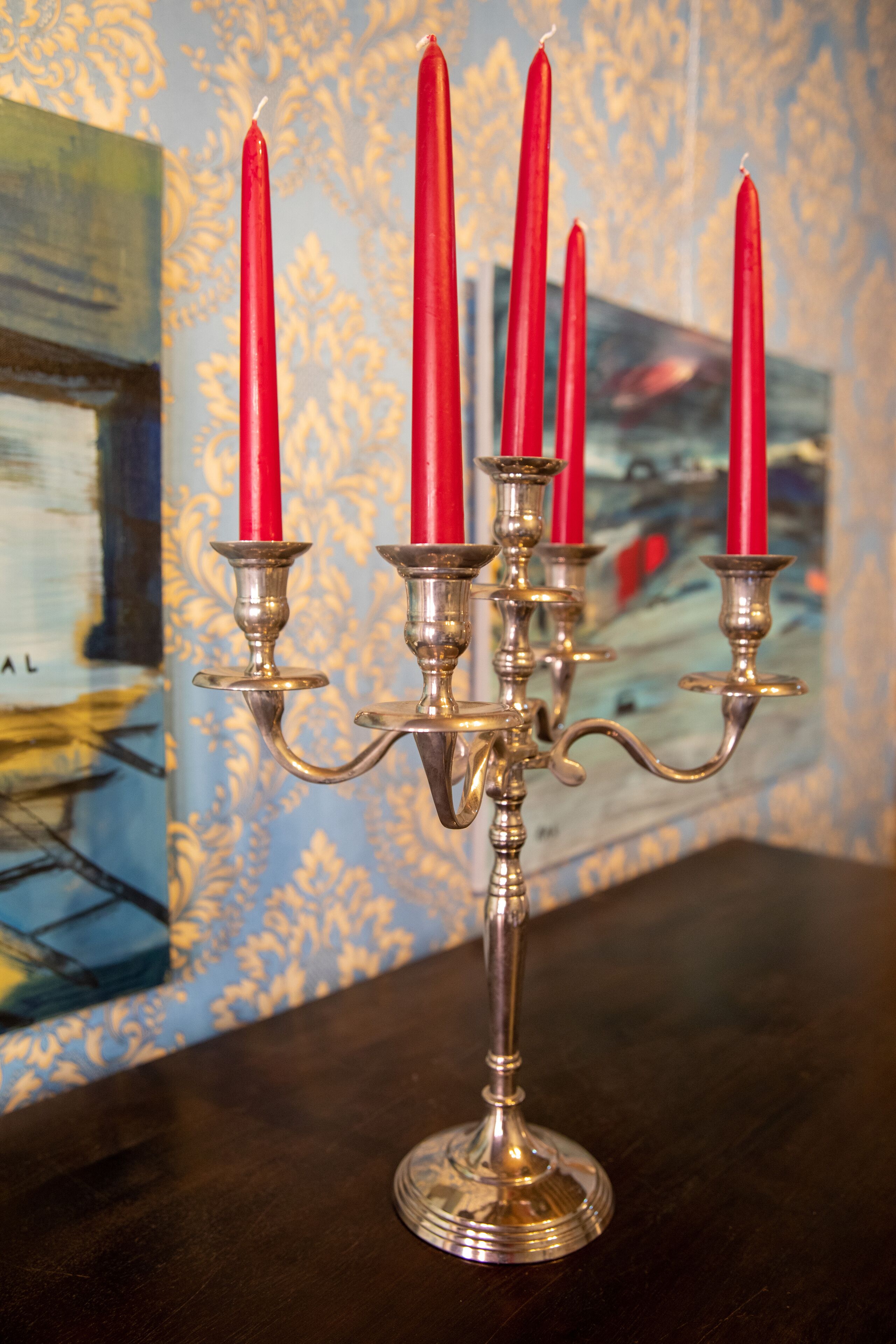 Un chandelier à quatre bougies rouges, posé sur une table en bois sombre, le tout sur fond d’un papier peint à motifs.