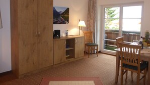 A room with a wooden cabinet, a desk with a lamp, a chair, and a dining table with chairs. There is a window with a view of greenery outside. - Ferienhotel Silberdistel garni (Bolsterlang)