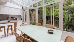 Een oranjerie met een eettafel en stoelen, omgeven door grote ramen met uitzicht op groen.
