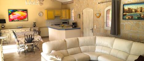Amplia sala de estar con un sofá blanco de cuero curvo, una mesa de centro de vidrio y una cocina con armarios amarillos y una pared de piedra.