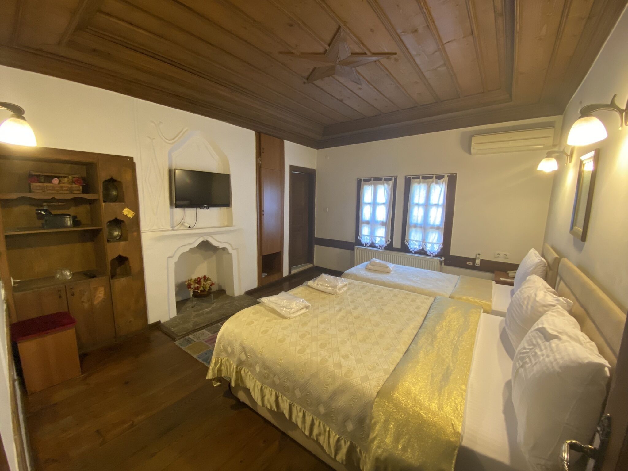 Habitació d'hotel amb sostre de fusta, una llar de foc, un llit amb roba de llit blanca, un televisor muntat a la paret i una finestra amb cortines.