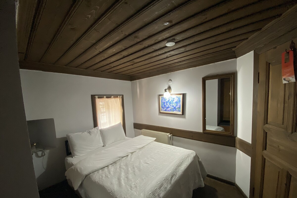 A bedroom with a wooden ceiling, a bed with white linens, a wall-mounted light, a framed picture, and a mirror.