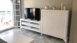 A modern living room with a flat-screen TV on a white entertainment unit, a white cabinet with diamond-patterned doors, and a large window with a view of the outdoors. - BALCON DEL MAR 2-3 A, APARTMENT ON THE BEACH FRONT (Mijas)