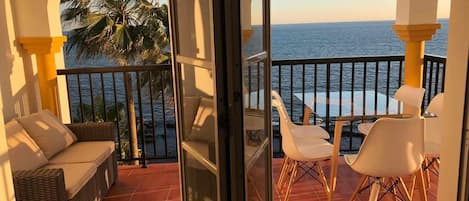 A balcony with a wicker sofa and white chairs, overlooking a palm tree and the sea.