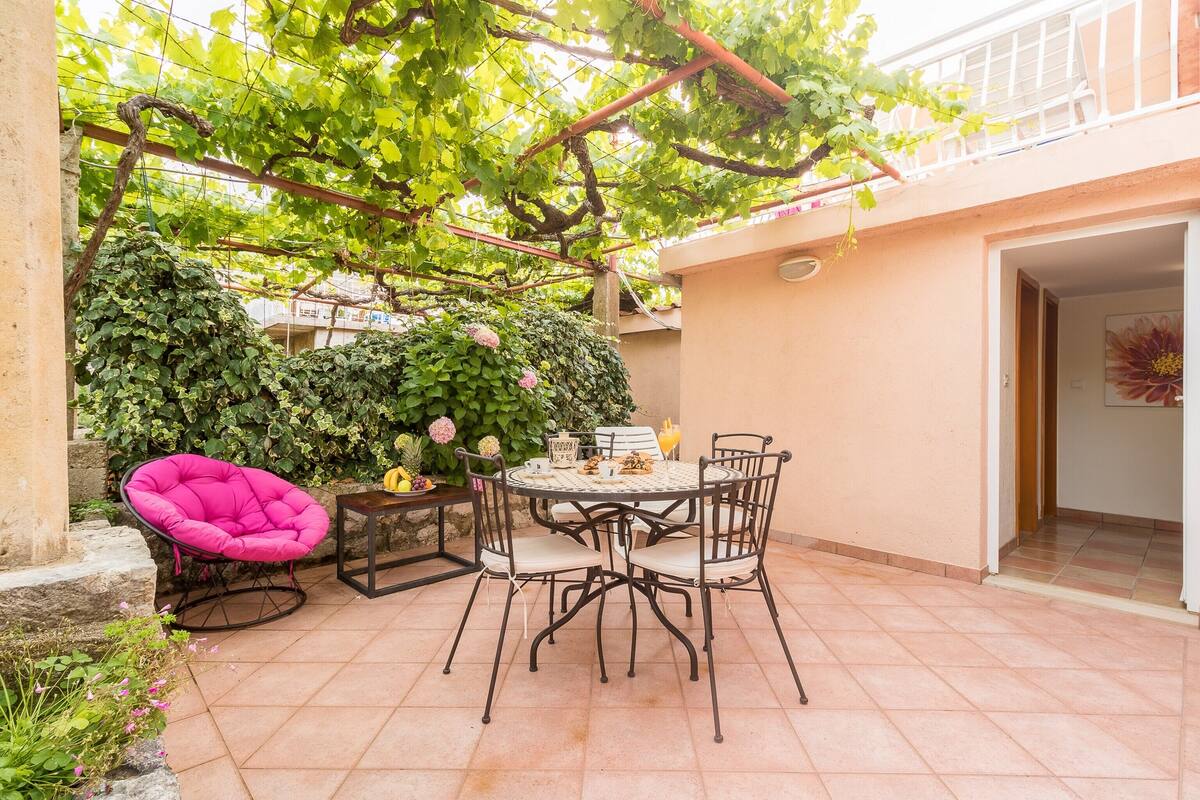 Un pati amb una taula rodona i cadires, envoltat per una paret verda i plena de vides.