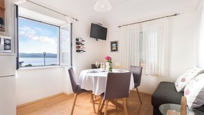 A dining area with a table set for two, a sofa, a TV mounted on the wall, and a window with a view of the sea. - Apartments Lotea (Konavle)