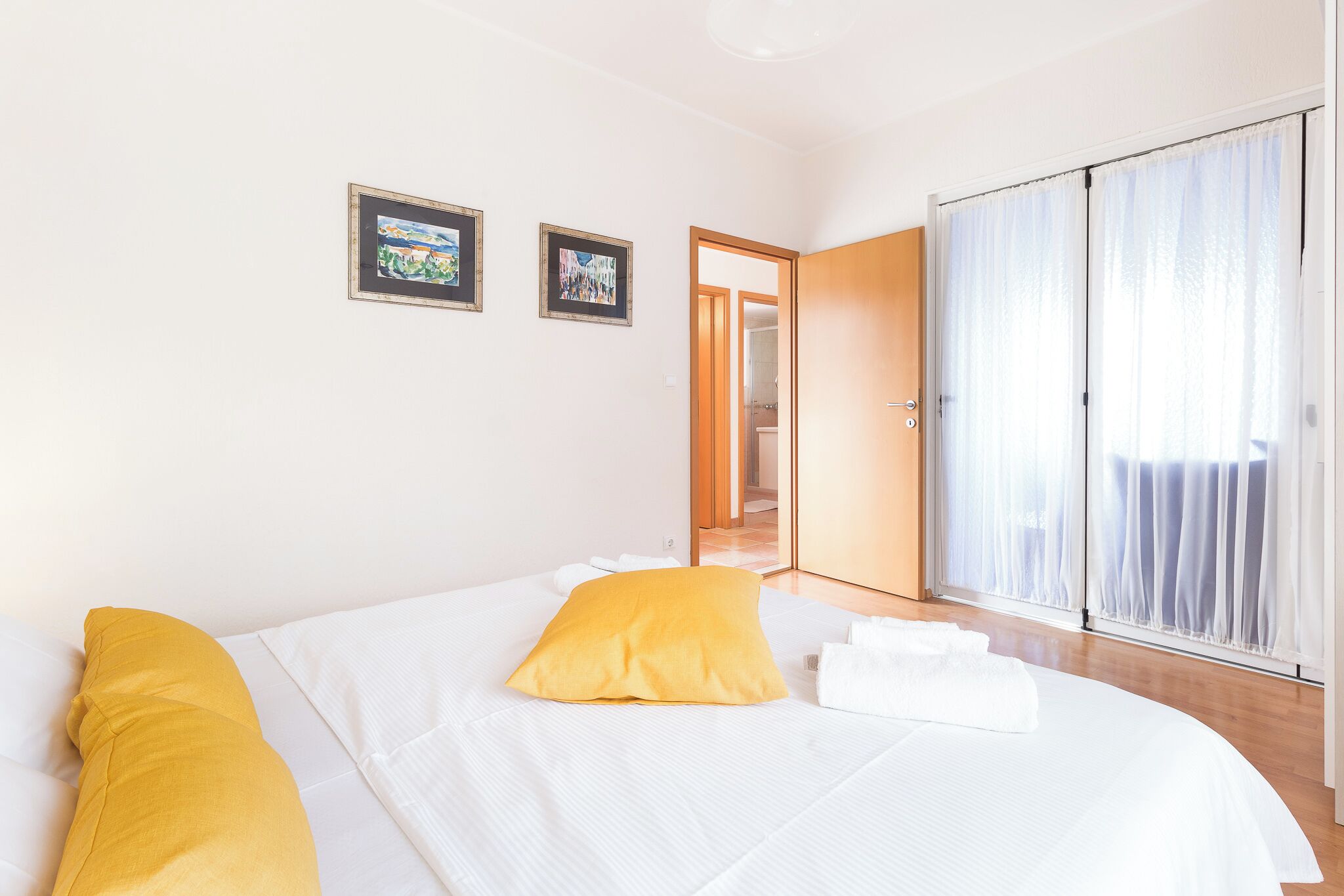 Ein Hotelzimmer mit einem Bett, einem gelben Kopfkissen, weißer Bettwäsche und einem großen Fenster mit durchsichtigen Vorhängen.