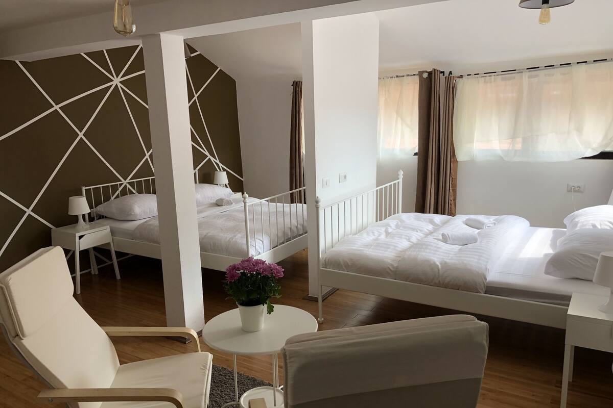 Ein Hotelzimmer mit zwei Betten, einem Sessel, einem kleinen Tisch mit Pflanze und einem Fenster mit Vorhängen.