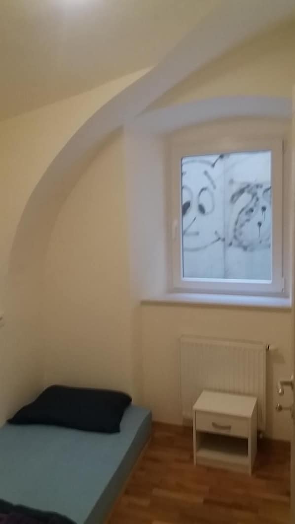 A small, single-room apartment with a sloped ceiling, a bed, a small table, a radiator, and a window with a view. - Central Antique Apartments (Ljubljana)