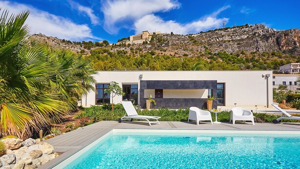 A modern pool area with clear blue water, white lounge chairs, and a view of a mountain with a building on top.
