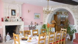 A dining room with a fireplace, wooden chairs, a table set for four, and a kitchen area visible through an archway. - Le Logis de Faugerit (Frontenay-Rohan-Rohan)