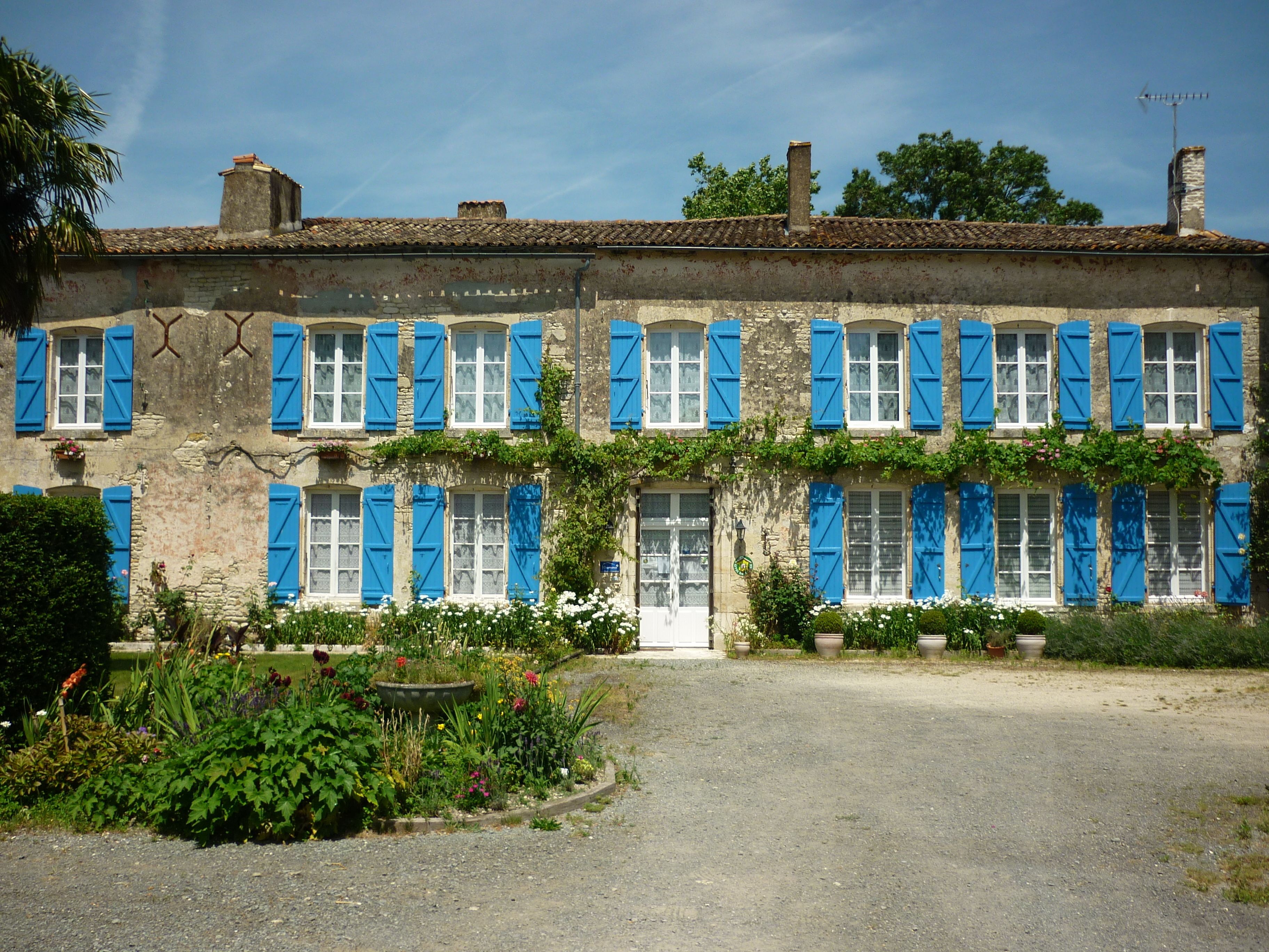 A two-story building with blue shutters, a gravel driveway, and a garden with various plants.