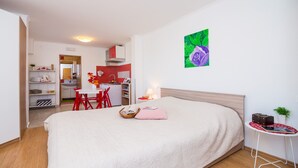 Quarto de hotel com cama, uma mesinha com abajur, uma cadeira vermelha, uma cozinha ao fundo e um quadro na parede.