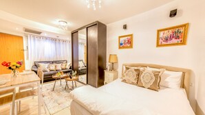 Habitación de hotel con una cama grande, un comedor con mesa y sillas, un sofá, una pequeña mesa redonda con una lámpara y dos cuadros en la pared.