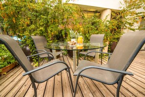 A glass table with four chairs, surrounded by lush greenery and a wooden deck.