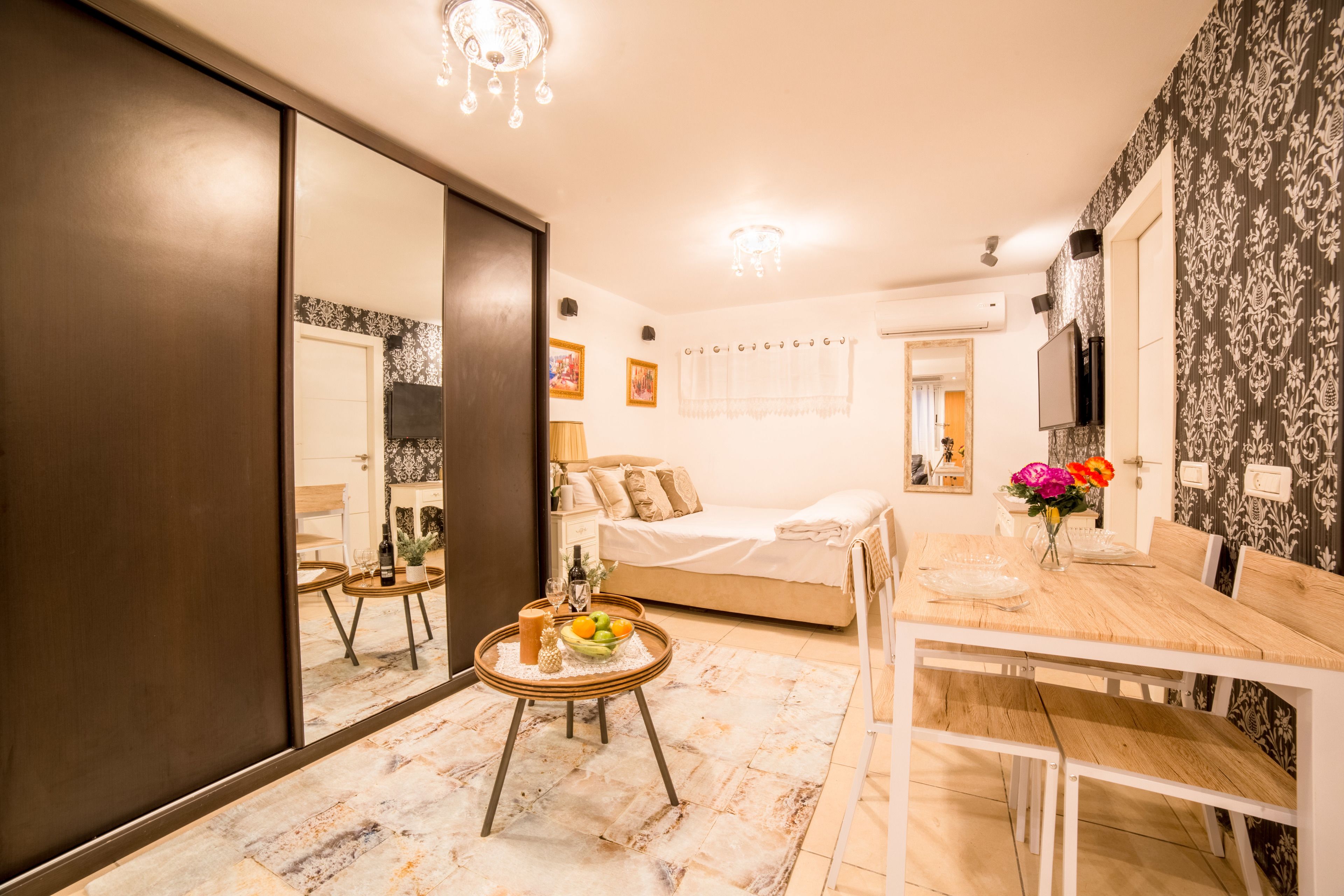 A bedroom with a bed, bedside table, and a mirror. There is a dining area with a table and chairs.