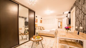 Un dormitorio con cama, mesita de noche y espejo. Hay una zona de comedor con mesa y sillas.