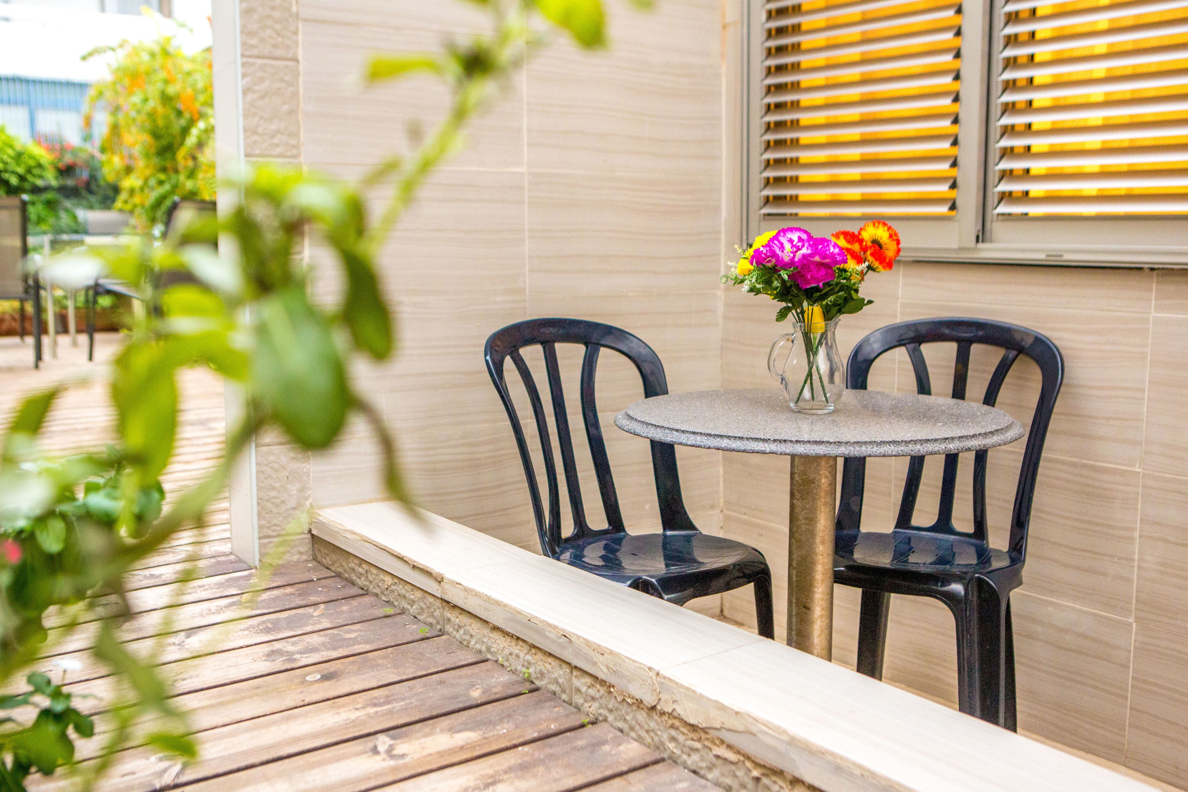 A patio area with a round table and two chairs, a vase with flowers, and a view of the outdoors.