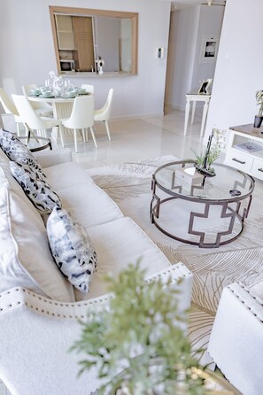 A modern living room with a white sofa, a round coffee table, and a dining area with white chairs.