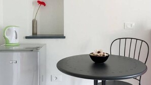 A compact kitchen area with a refrigerator, a small table with a bowl, a chair, and a vase with a red flower on a shelf. - Arbanella (Vernazza)