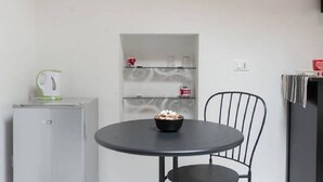 A small kitchen area with a round table and a chair, a refrigerator, and a shelf unit with various items. - Arbanella (Vernazza)