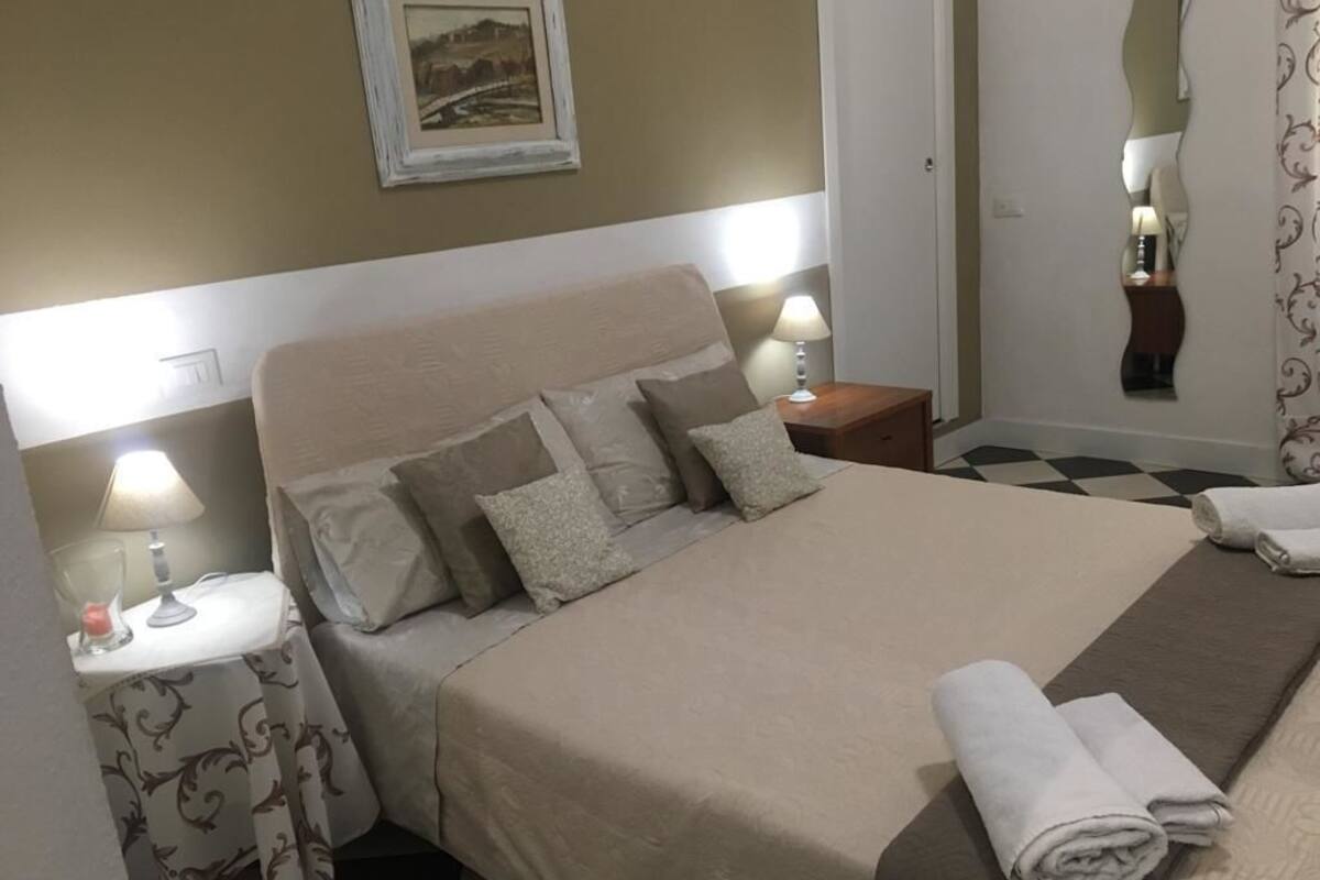 A neatly made bed with a beige bedspread and pillows, two bedside lamps, a framed picture on the wall, and a checkered floor.