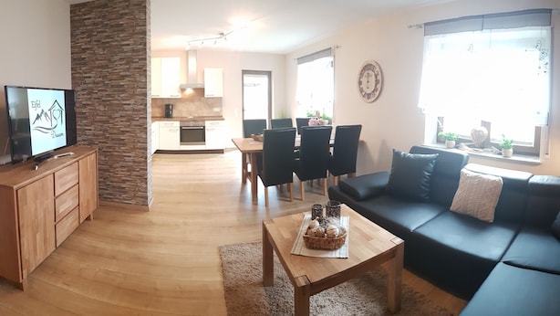 A modern living room with a black sofa, wooden coffee table, and a TV on a wooden cabinet. A kitchen is visible in the background with a dining area. - Eifel Dream Zingsheld (Fleringen)