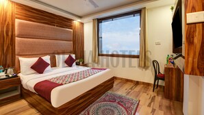 A hotel room with a large bed, wooden headboard, a ceiling fan, a window with curtains, a desk with a chair, and a small plant on the desk. - Olivia Hotels @ Delhi Airport (New Delhi)