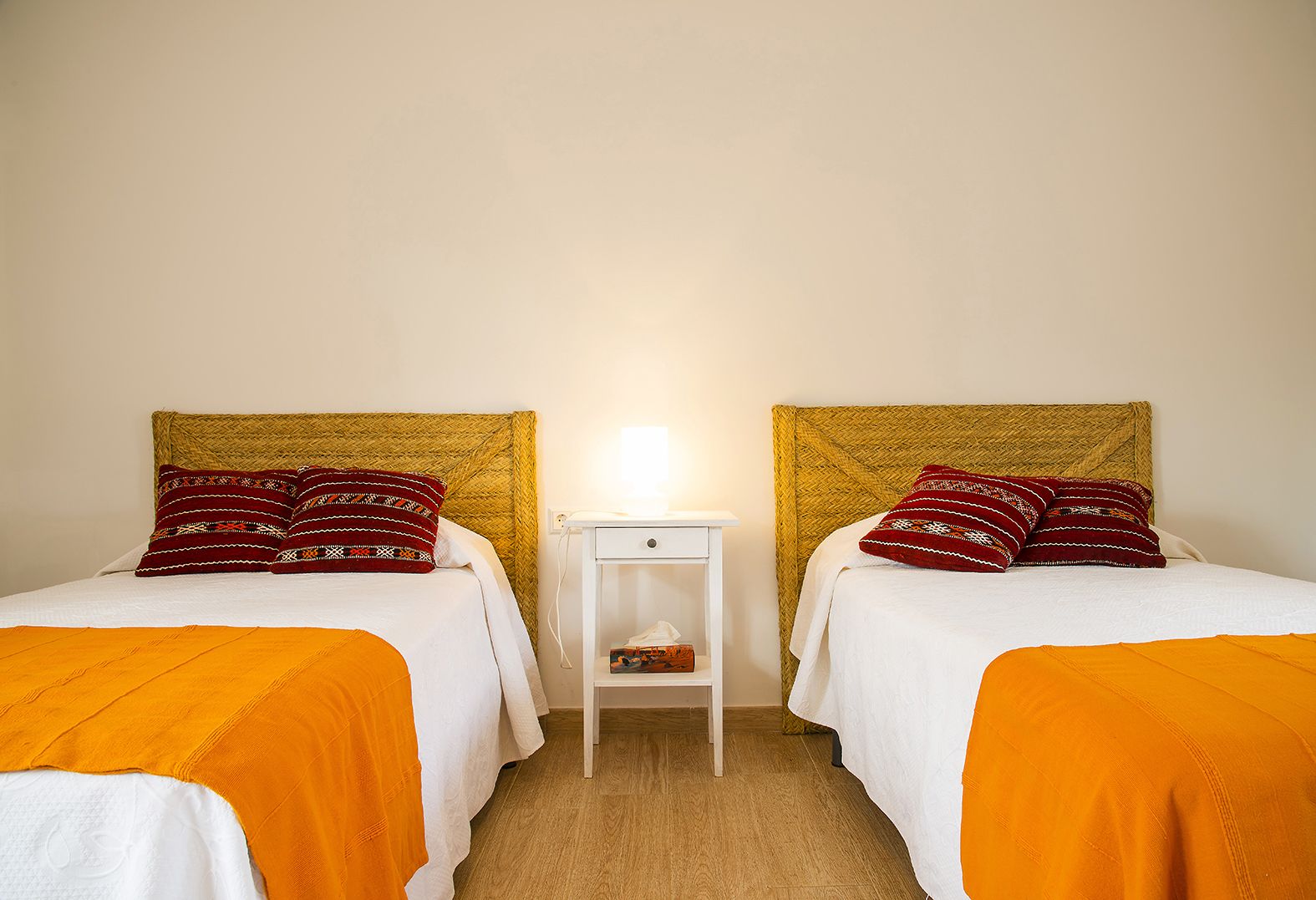 Una habitación con dos camas, cada una con un cabecero tejido, ropa de cama blanca y almohadas rojas con estampado. Hay una mesita de noche pequeña con una lámpara entre las camas.