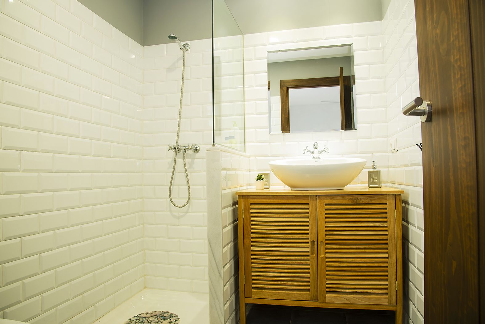Un baño con azulejos blancos tipo metro, un tocador de madera, un lavabo grande, un espejo y una ducha con cabezal de ducha manual.