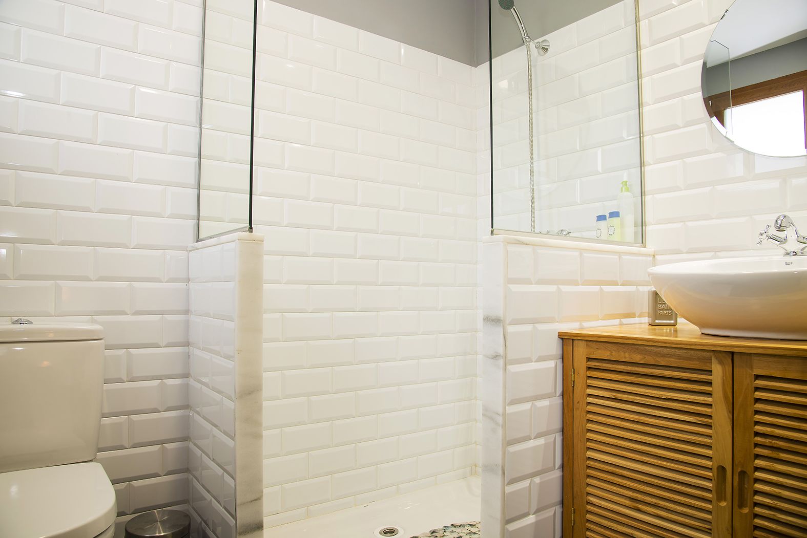 Un baño con azulejos blancos tipo metro, ducha con mampara de vidrio, tocador de madera y lavamanos blanco.