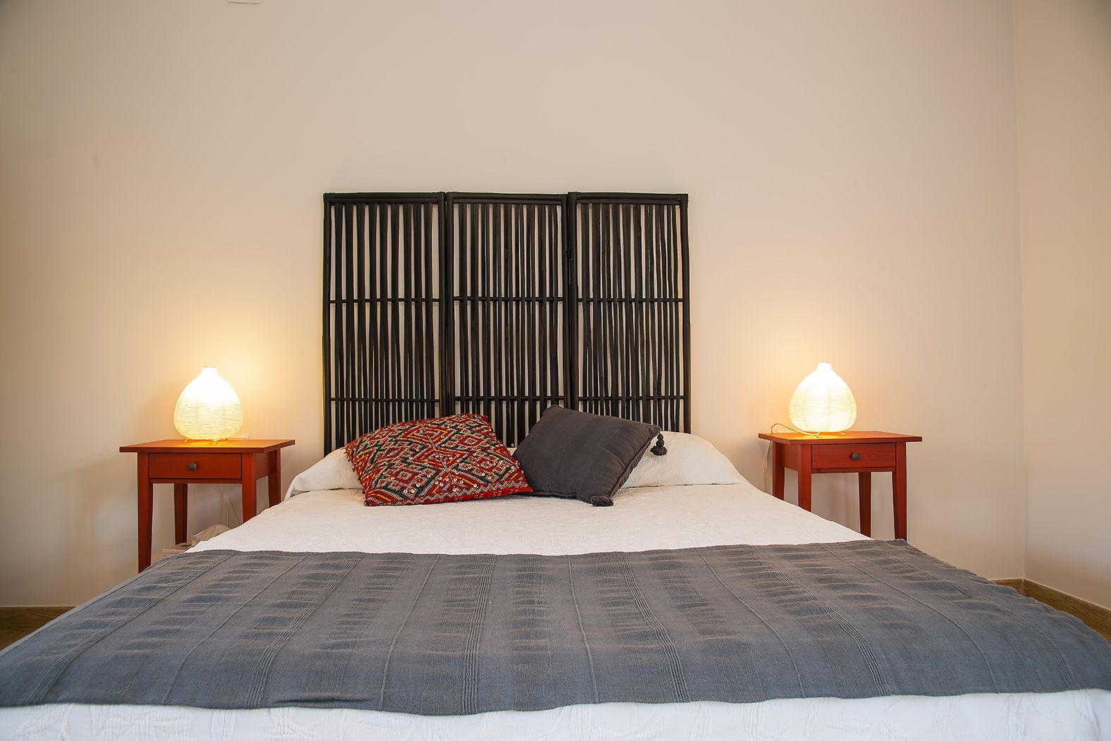 A neatly made bed with a grey blanket, two bedside tables with lamps, and a bamboo headboard.