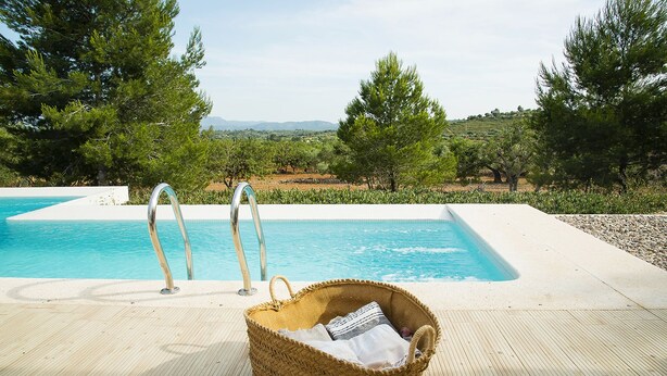 Zona de piscina con una cesta de mimbre que contiene una toalla y un libro, escaleras metálicas para la piscina, y vistas a árboles y colinas al fondo.