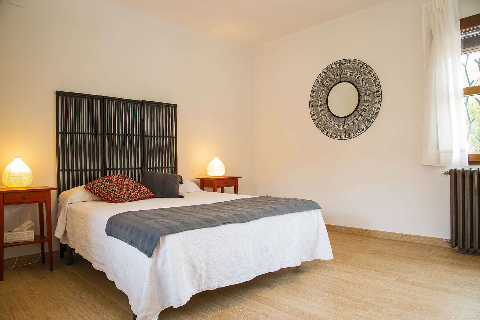 A bedroom with a bed, two bedside tables with lamps, a wall-mounted mirror, and a radiator.