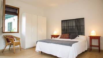 Een slaapkamer met een bed, twee nachtkastjes met lampen, een rieten stoel, een kledingkast en een spiegel aan de wand.