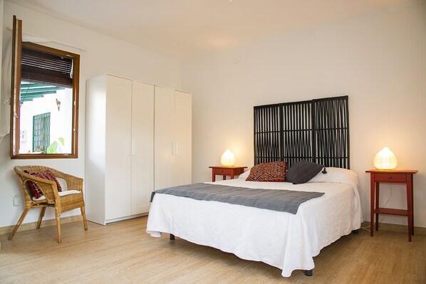 Un dormitorio con una cama, dos mesitas de noche con lámparas, un sillón de mimbre, un armario y un espejo adosado a la pared.