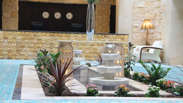 A reception area with a stone wall, a reception desk, a clock, and a fountain. - Saray Hotel (Amman)