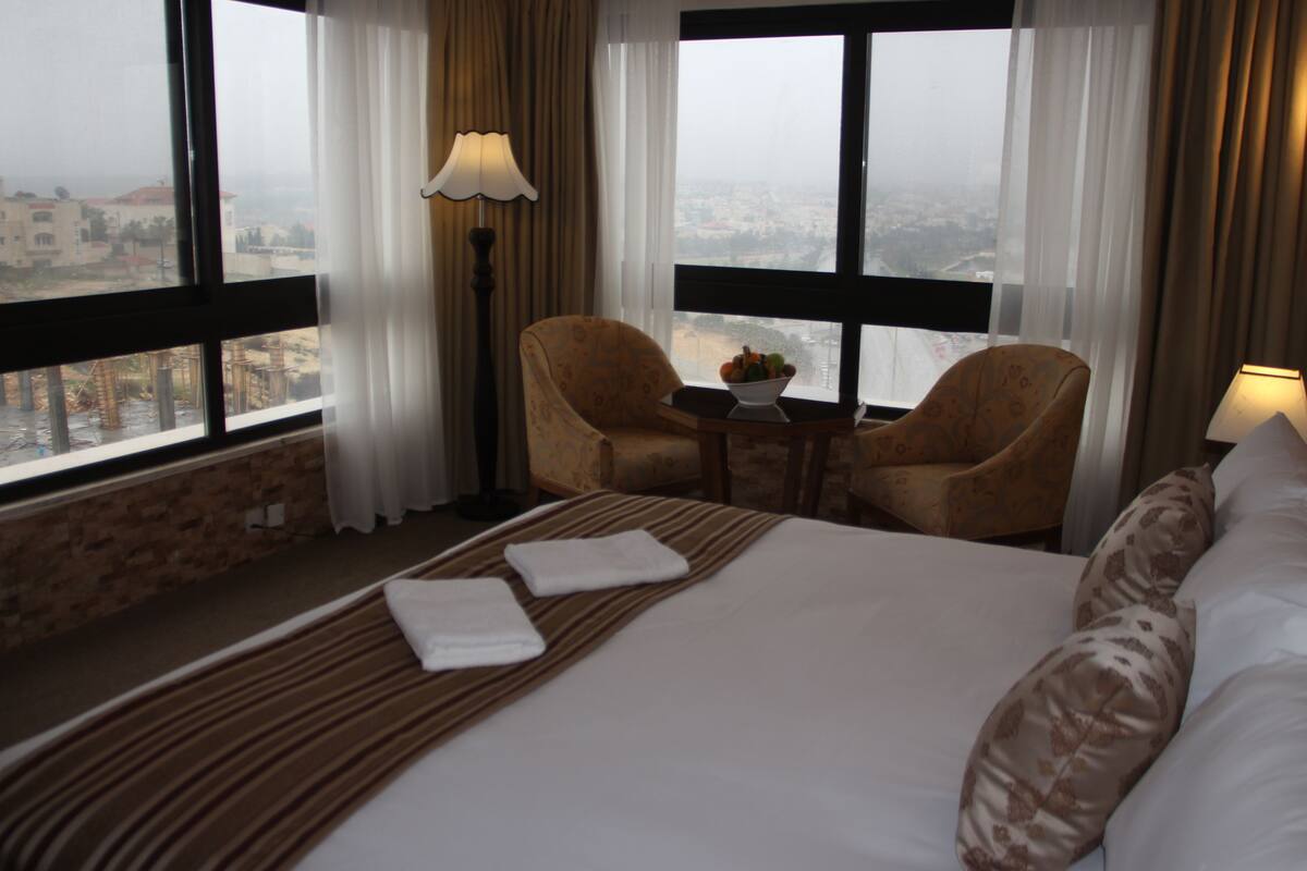 Habitación de hotel con una cama grande, dos sillones, una mesita con un frutero y vistas a la ciudad a través de amplios ventanales.