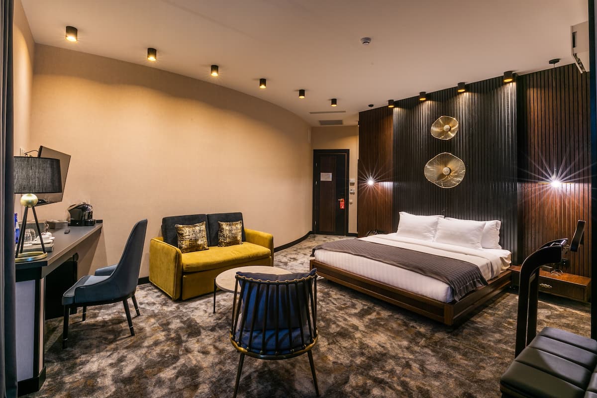 A hotel room with a bed, a desk with a lamp, a sofa, a chair, and a small round table.
