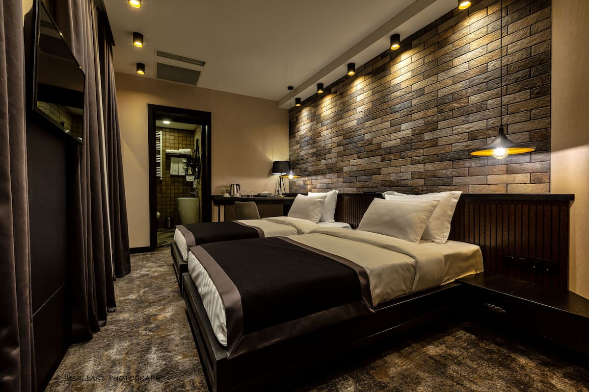 A hotel room with two beds, a brick-patterned wall, a desk with a lamp, and a view of a bathroom.